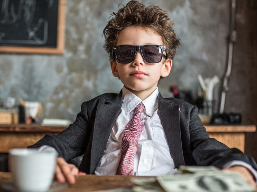 piores-conselhos-sobre-empreender-empreendedor-nato. Um menino com óculos escuros, vestido como um empresário, com terno e gravata, sentado em uma mesa, sobre a qual tem um xícara de café e notas de dinheiro